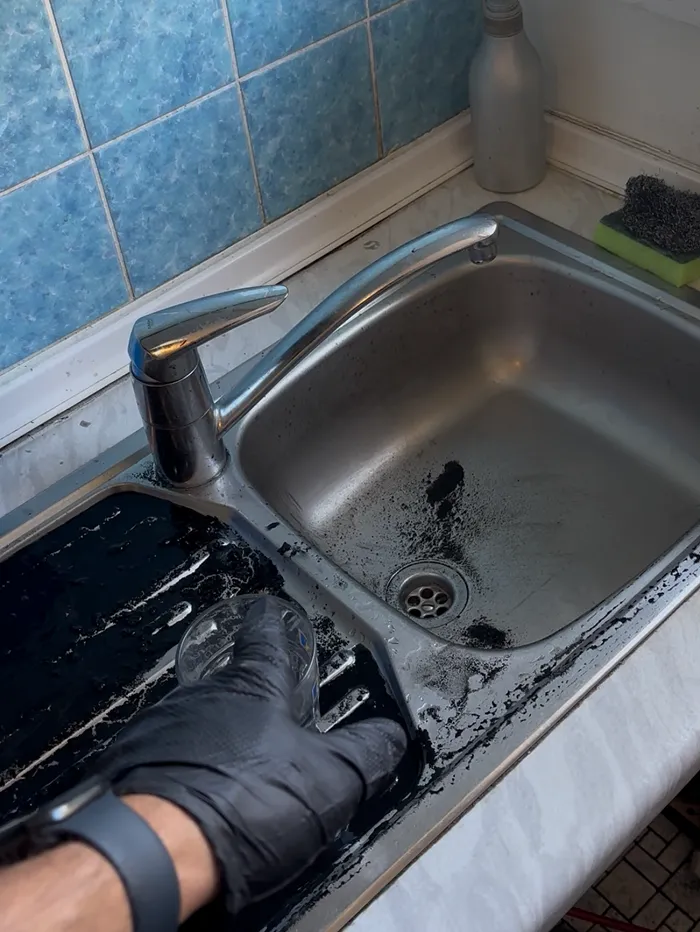 Une main gantée de noir tenant un verre sous le robinet d'un évier de cuisine, avec des traces noires autour de l'évier.